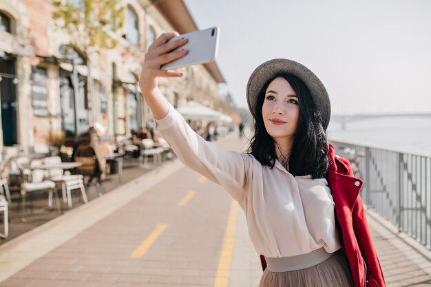 Mulher feliz de cabelos escuros em roupa romântica fazendo selfie perto do café da rua