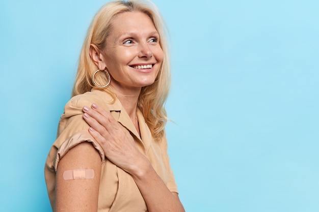 Mulher feliz de meia-idade é inoculada no ombro e vacinada na clínica mostra ombro com bandagewears vestido bege isolado na parede azul