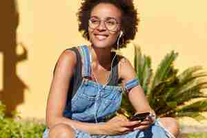 Foto grátis mulher feliz de pele escura com corte de cabelo afro, usa aplicativo no celular para ouvir música, gosta de som bom, usa macacão jeans, senta-se com as pernas cruzadas, modela sobre parede amarela com plantas tropicais