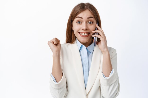 Mulher feliz do escritório, elegante senhora de terno branco, atender o telefonema e pular de felicidade, receber ótimas notícias, ganhar e comemorar, em pé sobre a parede branca