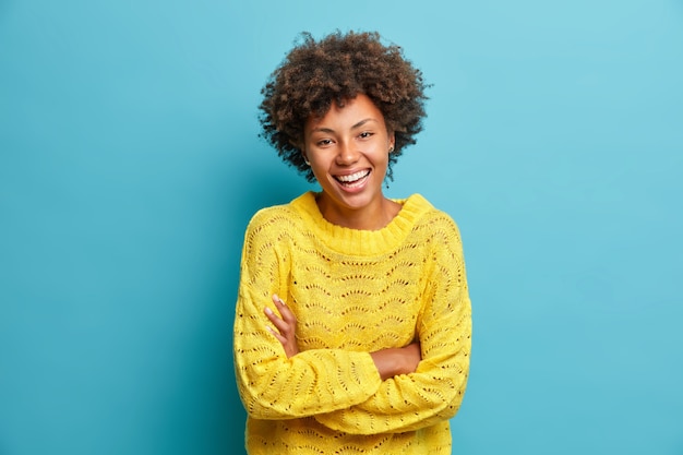 Mulher feliz e alegre ri, mantém os braços cruzados e expressa emoções positivas, sorrisos de felicidade, vestida com um macacão casual isolado na parede azul, se diverte ou ouve uma piada engraçada