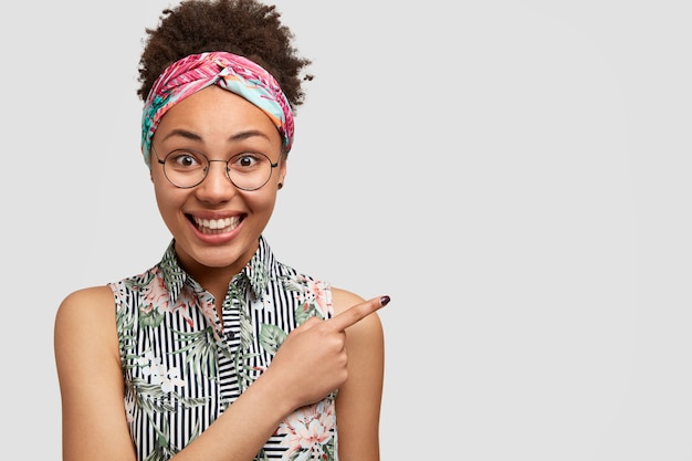 Foto grátis mulher feliz e animada em roupas elegantes indica à parte, como mostra algo sobre uma parede branca em branco, poses internas