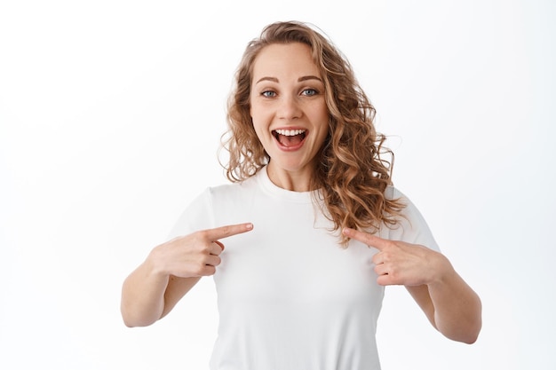 Mulher feliz e animada sorrindo, apontando para si mesma com rosto alegre e cândido, autopromoção, gesto escolha-me, em pé sobre fundo branco