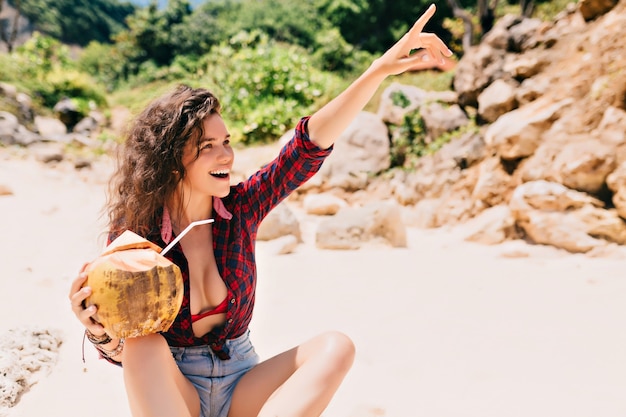 Mulher feliz e animada vestida de shorts e camisa brilhante sentada na praia com coquetel de coco e apontando para o lado