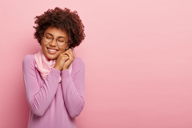 Foto grátis mulher feliz e atraente com penteado afro, sorri com alegria, expressa positividade, mantém os olhos fechados de prazer, tem sorriso agradável, usa óculos e suéter transparentes, modelos de interior sobre parede rosada