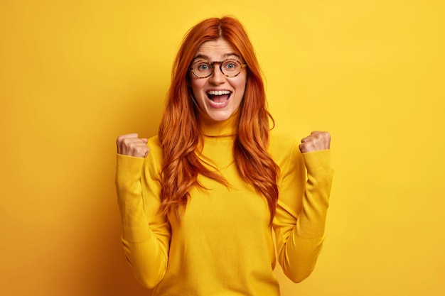 Mulher feliz e cheia de alegria se sente como se a vencedora festejasse algo levanta os punhos e exclamava sim, faz o gesto do vencedor alegrar-se com a vitória usando uma blusa casual de gola alta.