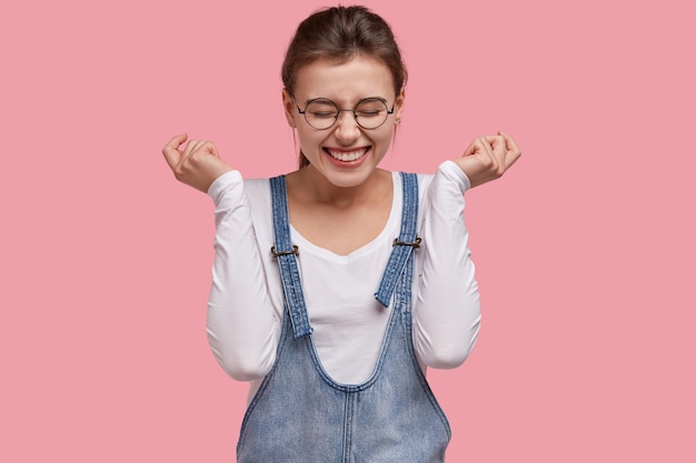 Mulher feliz e contente ri de uma anedota engraçada, abre os punhos, tem um sorriso cheio de dentes, usa óculos redondos e macacão jeans, modelos sobre fundo rosa