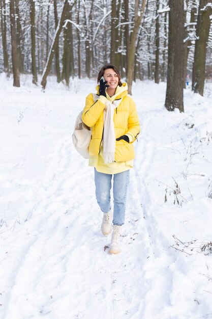 Mulher feliz e de ótimo humor caminha pela floresta de neve de inverno e conversa animadamente ao telefone, aproveitando o tempo ao ar livre no parque