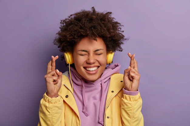 Mulher feliz e desejosa com cabelo crespo natural antecipa prazer e boas notícias, cruza os dedos e sorri amplamente, espera pela realização de um sonho, usa fones de ouvido estéreo, ouve música agradável.