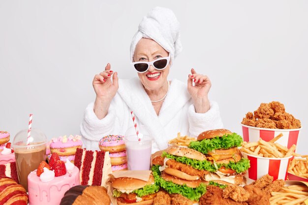 Mulher feliz e enrugada e despreocupada, sorrindo alegremente com óculos de sol, curtindo a refeição do dia rodeada de junk food