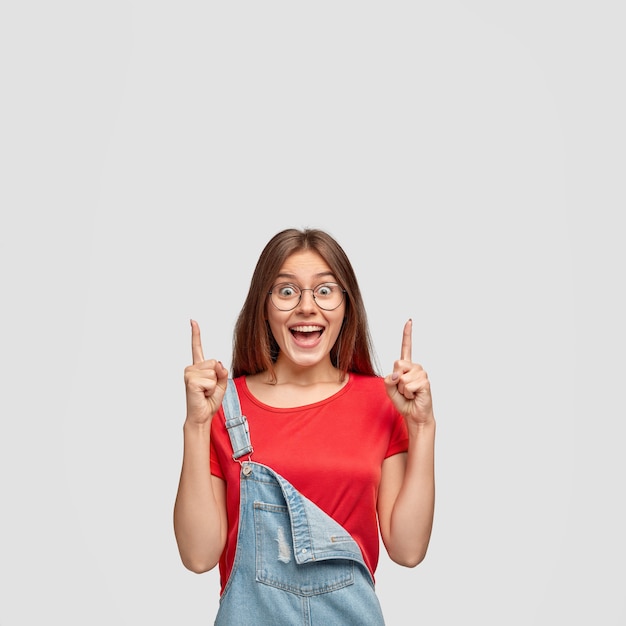 Foto grátis mulher feliz e feliz com expressão satisfeita, aponta com os dois dedos indicadores para cima, fascinada por algo