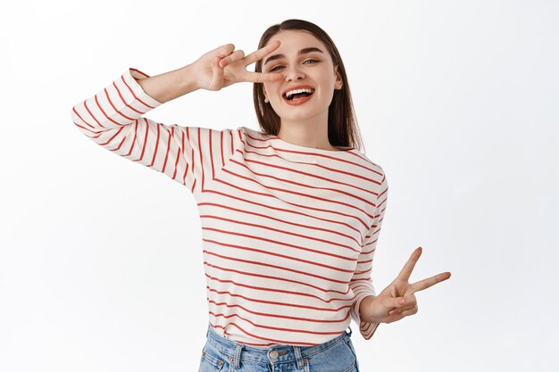 Mulher feliz e positiva, dançando com os sinais V da paz e rindo alegre, aproveitando fins de semana de lazer, posando alegre contra um fundo branco