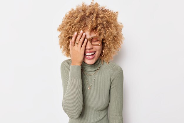 Mulher feliz e positiva faz sorrisos da palma do rosto com alegria, mantém os olhos fechados, expressa emoções positivas autênticas usa óculos e gola olímpica isolada sobre fundo branco. Conceito de pessoas e alegria