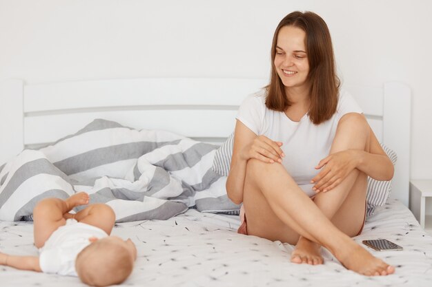 Mulher feliz e positiva vestindo camiseta branca e shorts, sentado na cama coberta de lençol branco com sua filha infantil, mulher de cabelos escuros, olhando com um sorriso para seu adorável filho.