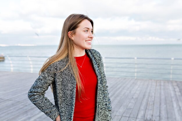 Mulher feliz e sorridente de cabelos compridos com grandes olhos azuis, camisa vermelha e casaco cinza caminhando perto do mar