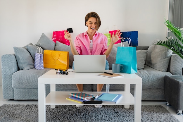 Mulher feliz e sorridente em uma camisa rosa no sofá em casa entre sacolas coloridas segurando um cartão de crédito e pagando online no laptop