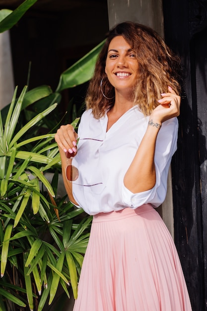 Mulher feliz e tranquila com cabelo curto e encaracolado em saia longa rosa e camisa branca sozinha fora de sua villa