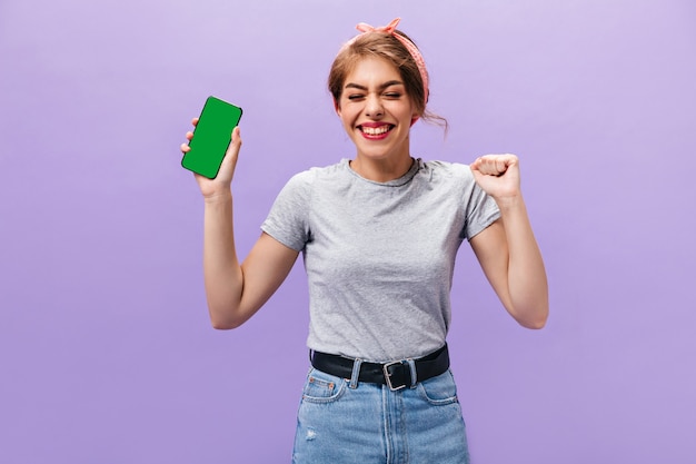 Mulher feliz em camisa cinza segura o telefone. Menina alegre com bandana rosa na saia jeans com faixa preta se diverte no fundo isolado.