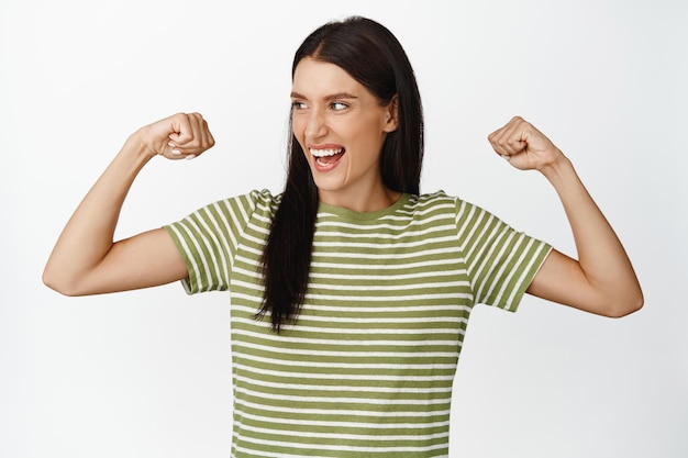 Mulher feliz empoderada se sentindo forte flexionando bíceps mostrando seus músculos nos braços e rindo em pé sobre fundo branco