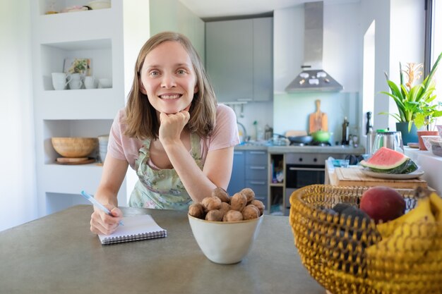 Mulher feliz encostado no balcão com frutas e nozes na cozinha, escrevendo notas no caderno e olhando para a câmera. Cozinhar em casa conceito