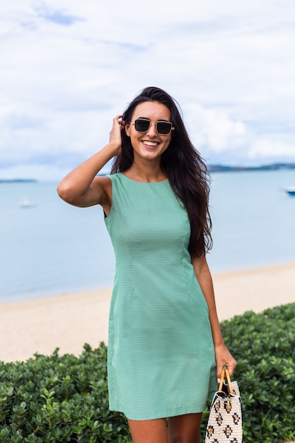 Mulher feliz muito elegante com vestido verde de verão com bolsa, óculos de sol nas férias, mar azul no fundo