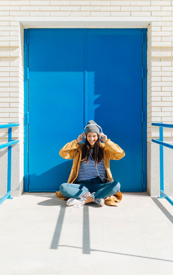 Foto grátis mulher feliz ouvindo música no telefone inteligente sentado contra a porta azul ao ar livre