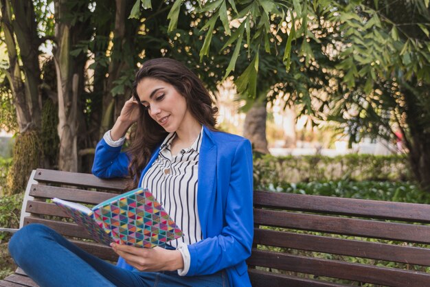 Foto grátis mulher feliz que lê um caderno em um banco de parque