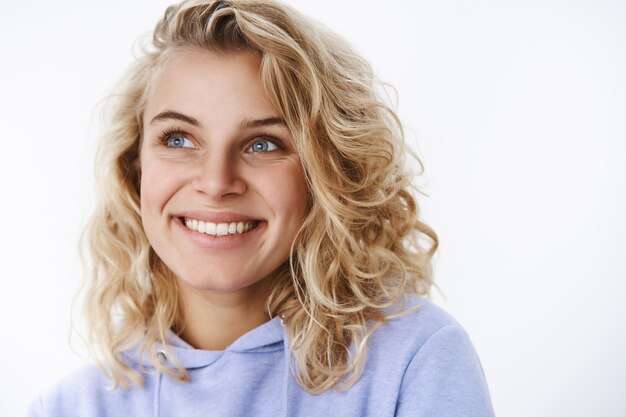 Mulher feliz relembrando memórias agradáveis, calorosas e ternas, sorrindo encantada olhando com belos olhos azuis no canto superior esquerdo enquanto lembra de um lindo momento sobre a parede branca