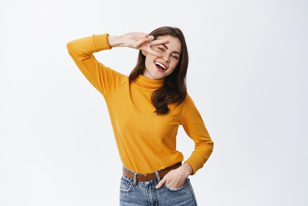 Mulher feliz rindo e mostrando vsign sobre o olho Garota sorrindo e fazendo gesto de paz em pé despreocupado contra o fundo branco