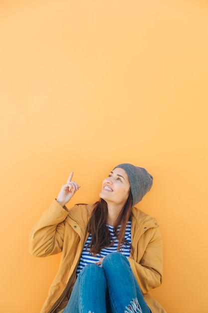 Foto grátis mulher feliz sentado contra o pano de fundo amarelo apontando para cima