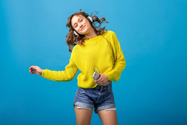 Mulher feliz sorridente atraente dançando ouvindo música em fones de ouvido em roupa elegante hippie isolada no fundo azul do estúdio, vestindo shorts e suéter amarelo