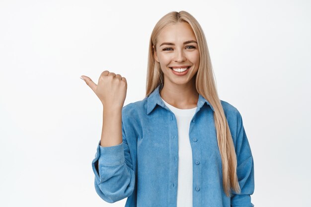 Mulher feliz sorridente com corte de cabelo loiro reto, apontando o dedo para a esquerda e parecendo confiante, aparecendo, anúncio de venda em branco.