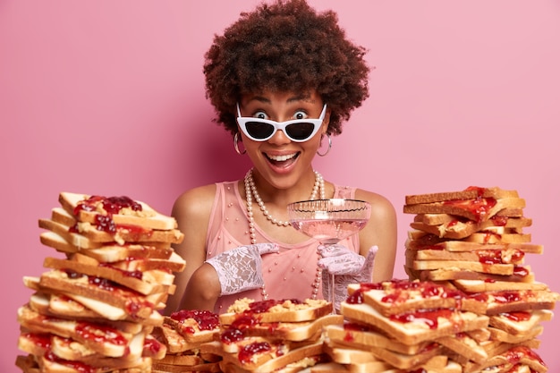 Foto grátis mulher feliz surpresa aponta para si mesma e não consegue acreditar no seu sucesso, usa óculos escuros e roupas elegantes, posa sobre a parede rosada de um café, rodeada de sanduíches, vai almoçar saboroso