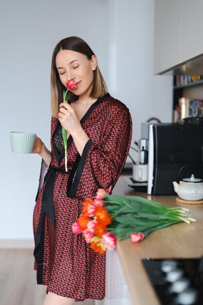 Mulher feliz tomando um café em casa na cozinha vestindo um roupão de seda enquanto desfruta de flores frescas