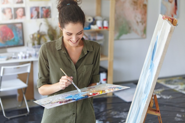 Mulher feliz trabalhando como pintor, em pé perto de cavalete, segurando o pincel, criando imagens abstratas com óleos coloridos, tendo bom humor e inspiração. Desenho feminino sobre tela. Conceito de arte