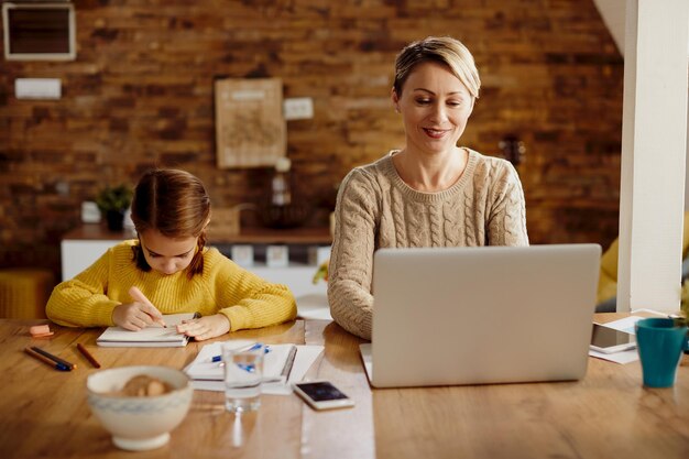 Mulher feliz trabalhando no laptop enquanto ensinava em casa sua filha pequena