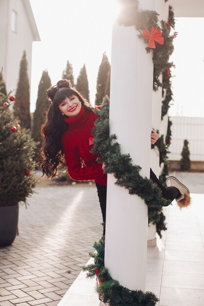 Mulher feliz vestindo um suéter vermelho e espiando por trás de uma coluna branca decorada com enfeites de Natal