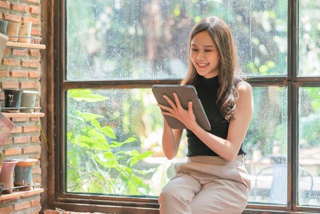 Mulher feminina casual da ásia relaxa a mão usando tablet internet surf escrevendo diário alegre sorrindo enquanto está sentado no interior quente do café perto de uma grande janela mulher asiática sente-se no banquinho trabalho casual no coffeeshop