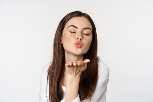 Mulher feminina mandando beijo aéreo na câmera, pose de paquera coquete, beijando, de pé sobre fundo branco. Copiar espaço