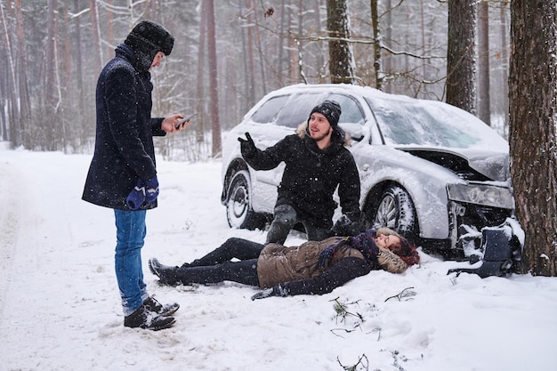 Mulher ferida está deitada na neve após o acidente de carro, o homem está tentando ajudá-la, o segundo homem está chamando a ambulância.