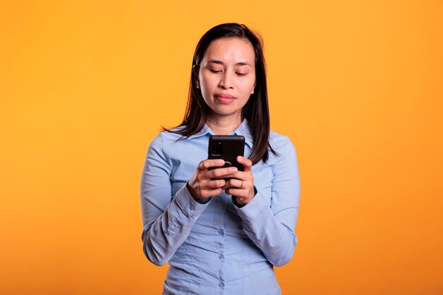 Foto grátis mulher filipina segurando mensagens de digitação no celular, conversando com um amigo remoto no estúdio sobre fundo amarelo. jovem adulto sorridente e alegre, aproveitando a discussão por sms durante o intervalo