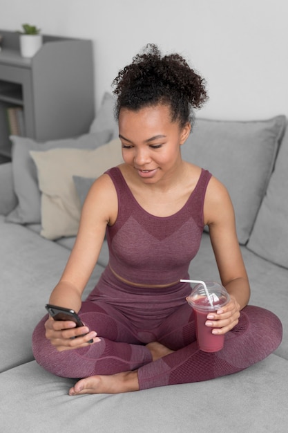 Mulher fitness tomando um suco desintoxicante enquanto usa um smartphone