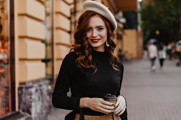 Mulher francesa tímida com cabelos longos, posando ao ar livre. Adorável senhora ruiva em pé na rua com uma xícara de café.