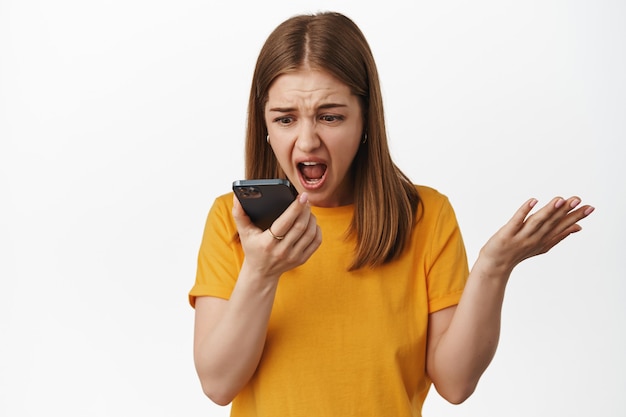 Mulher furiosa gritando com mensagem de voz de smartphone, fala no viva-voz e gritando com celular dinâmico, apertando as mãos frustrado, em pé contra uma parede branca.
