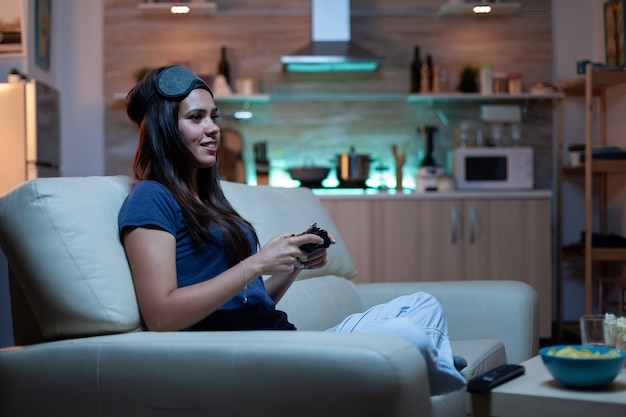 Mulher gamer se divertindo em casa sentada no sofá jogando videogame tarde da noite usando máscara para os olhos na testa