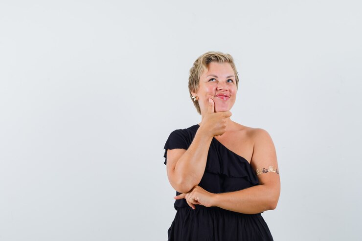 Foto grátis mulher glamorosa segurando a mão no queixo enquanto olha para cima com uma blusa preta e parece confiante. vista frontal. espaço para texto