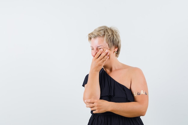 Mulher glamourosa, segurando a mão na boca de blusa preta e parecendo alegre. vista frontal. espaço para texto