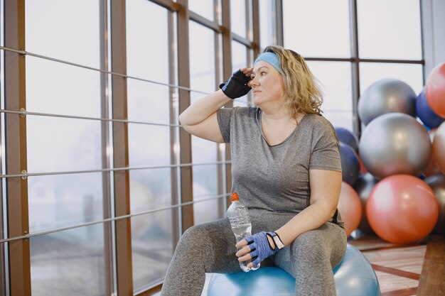 Mulher gorda fazendo dieta, fitness. Senhora sentada em um fitball e bebendo água.