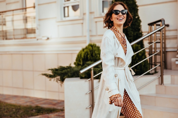 Foto grátis mulher graciosa de jaleco branco e óculos de sol, expressando emoções felizes. foto ao ar livre da bela senhora com roupa de outono elegante.