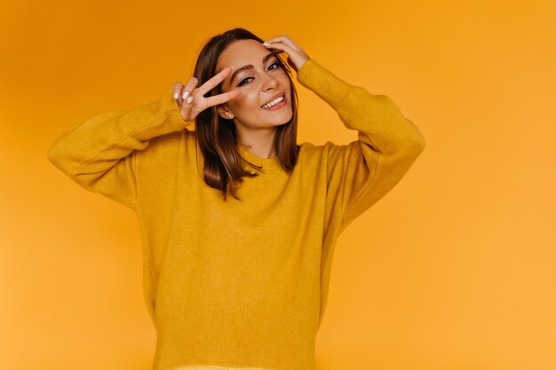 Mulher graciosa de suéter posando com o símbolo da paz. Foto interna de uma jovem refinada se divertindo na parede laranja.